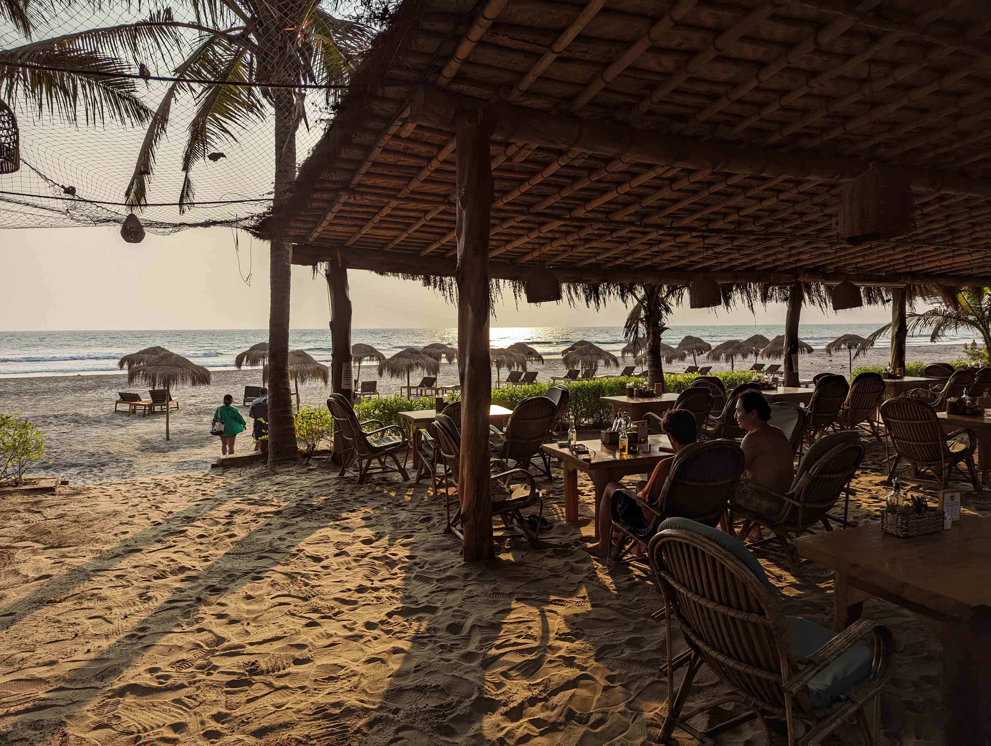 View of beach at Babu Beach Resort in North Goa with people enjoying their time at tables under a thatched roof and loungers with umbrellas.