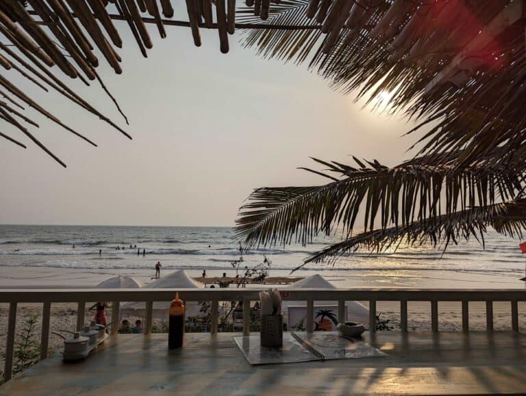 View of the beach in Goa from a table at a shack with the sun setting beyond palm tree fronds in the thatched roof and people in the distance on the beach.