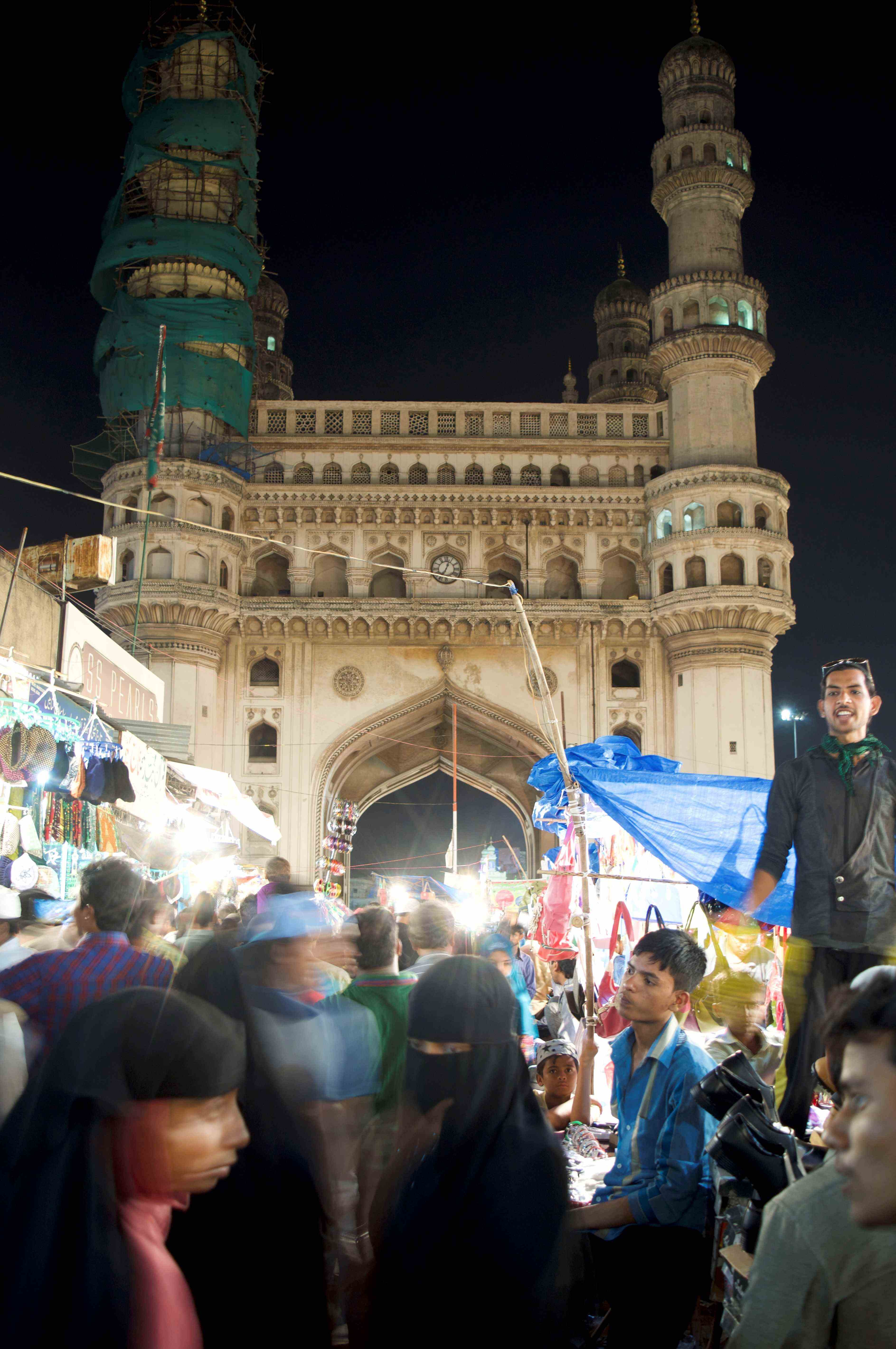 A crowded street market at night near the Charminar monument in Hyderabad, India. The illuminated Charminar, partially under scaffolding, serves as a stunning backdrop, while the bustling scene below features vendors selling various items under bright lights. People, including women wearing burqas and others in casual clothing, are moving through the market, adding to the lively atmosphere. A man stands elevated, possibly a vendor, engaging with the crowd. The scene reflects the cultural richness and energy of a typical evening in the city.