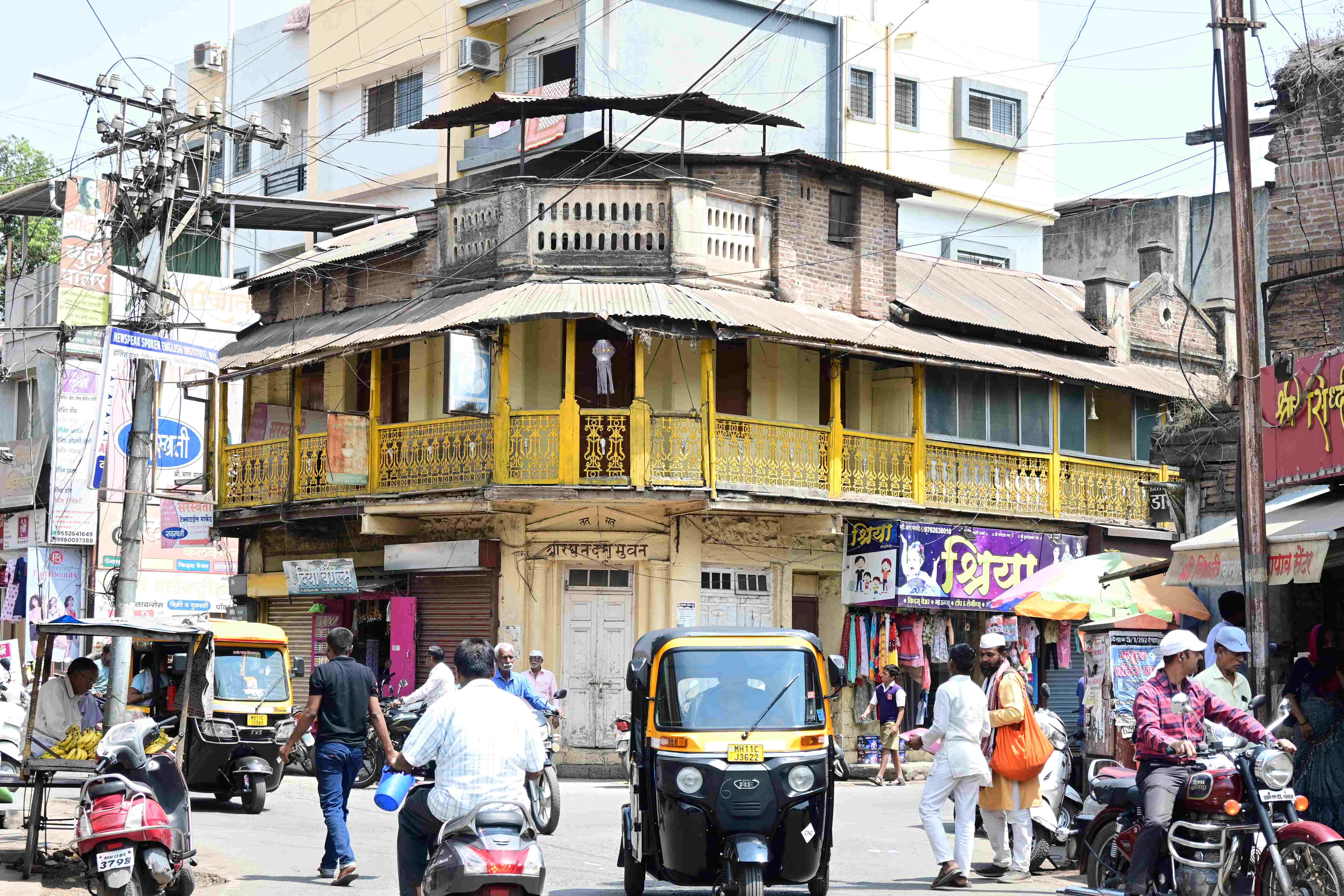 A busy street scene in an Indian city. The focal point is an old two-story building with a yellow balcony and ornate railings. The street is crowded with people, auto-rickshaws, motorcycles, and pedestrians. Colorful shop signs in Hindi and English line the buildings. Overhead, a tangle of electrical wires crisscrosses the sky. The scene captures the chaotic energy of urban life in India.