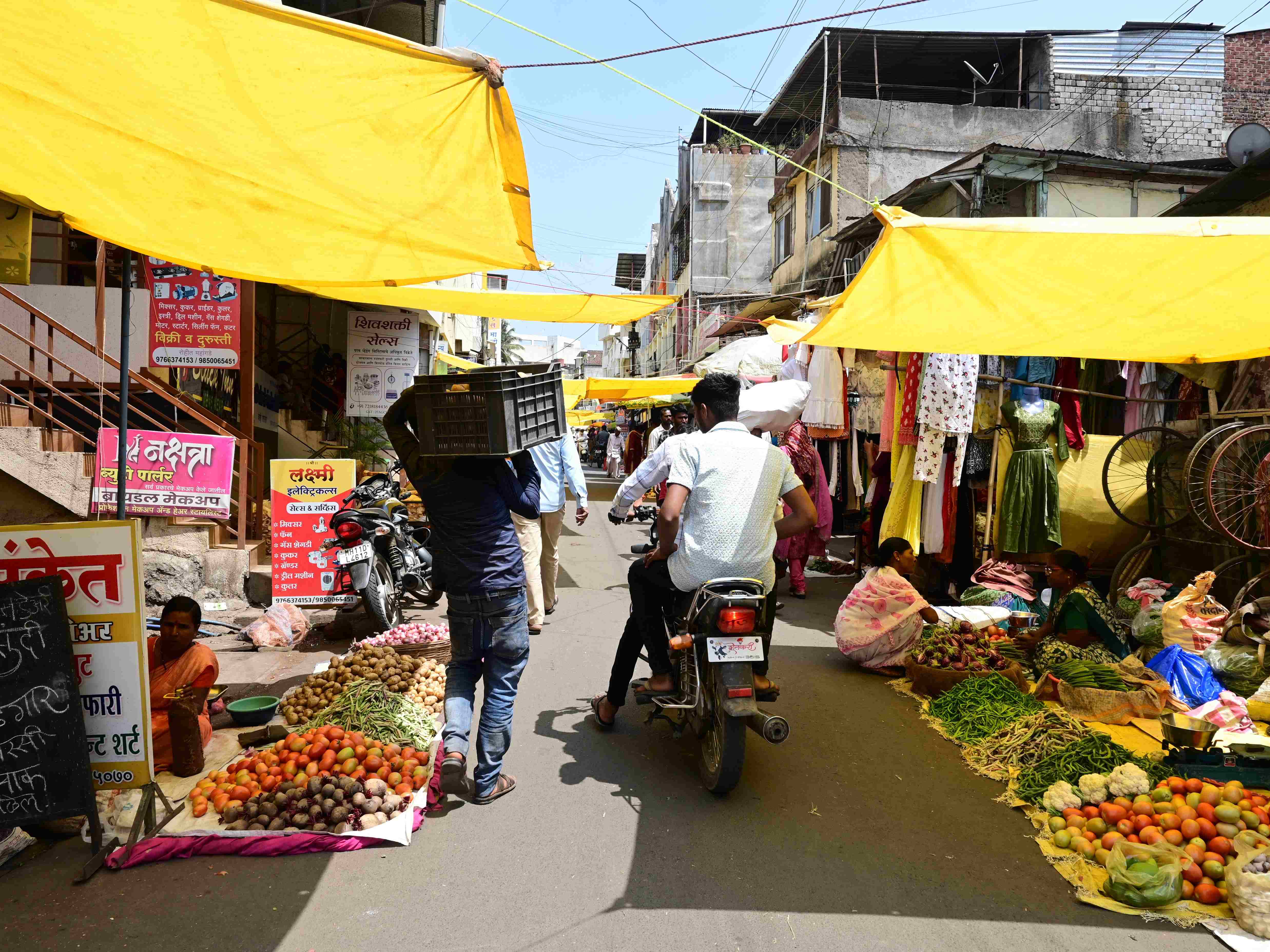 A street farmer's market in a village in India with fruit and vegetables lining the road, with yellow tent coverings, and two men on a motorcycle and a man carrying a large basket in the middle.