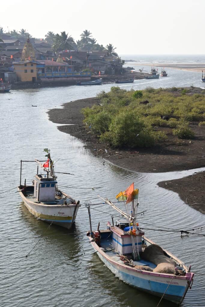 A picture of a coastal Indian village in Maharashtra, India. There are two boats in the foreground with a river flowing into the sea in the background. The village is in the background with an orange temple as the most prominent feature. 