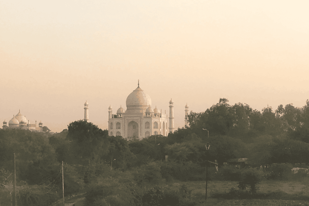 A distant view of the Taj Mahal at sunset, with its iconic white marble domes as softly glowing in the dusk light. The foreground includes fields and a few small structures, near Mehtab Bagh gardens.