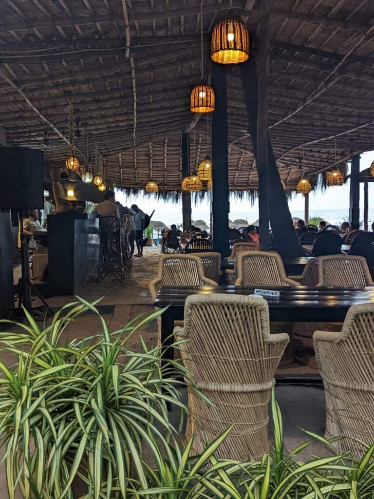 Inside Palm Grove shack on Ashvem Beach with thatched chairs at tables, hanging lights, and a roof made from bamboo and palm fronds. 