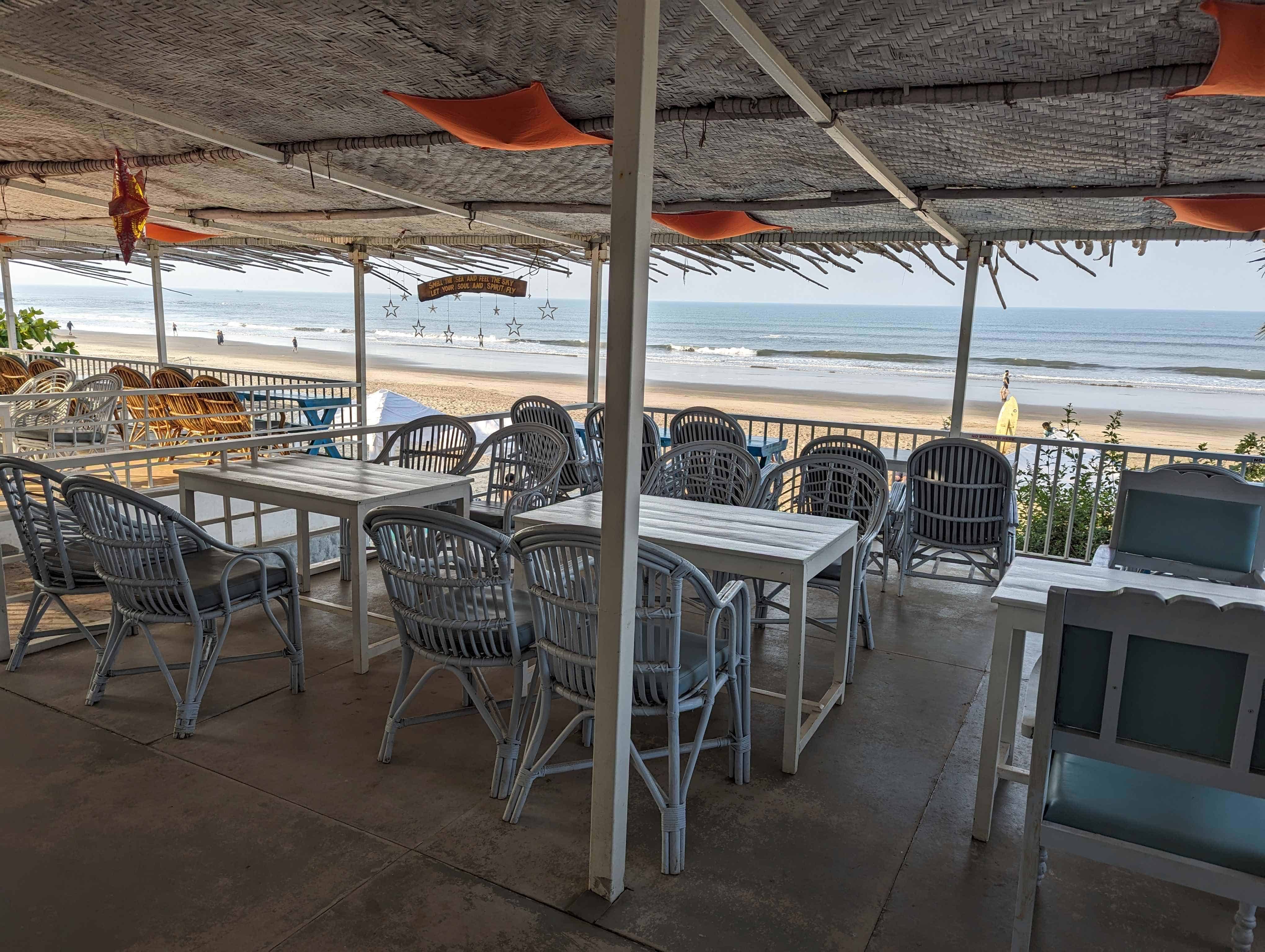 View of white tables and chairs under a thatched roof at S2 Beach Shack with Morjim Beach in the background.