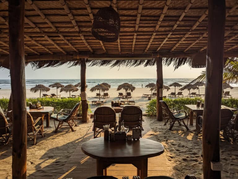 View of the beach in Goa from a beach shack restaurant with the sun setting beyond beach beds and the thatched roof.