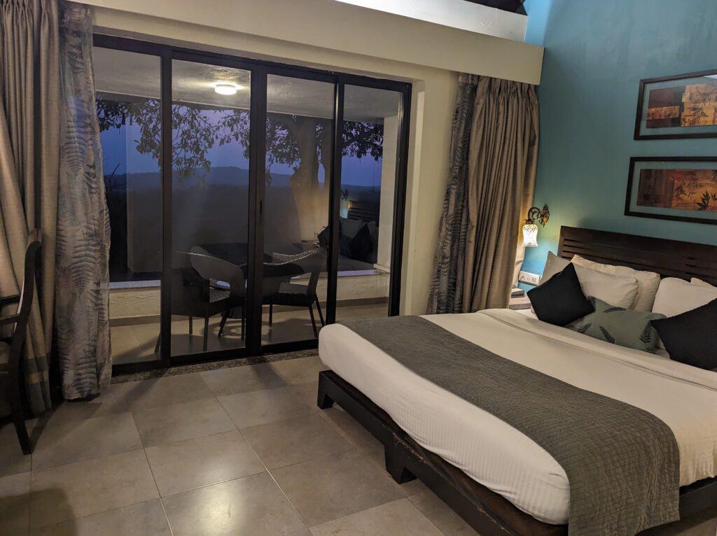 A view of the hotel room at Fern Dapoli showing a nice white bed, a tile floor, and a glass balcony with table and chairs.