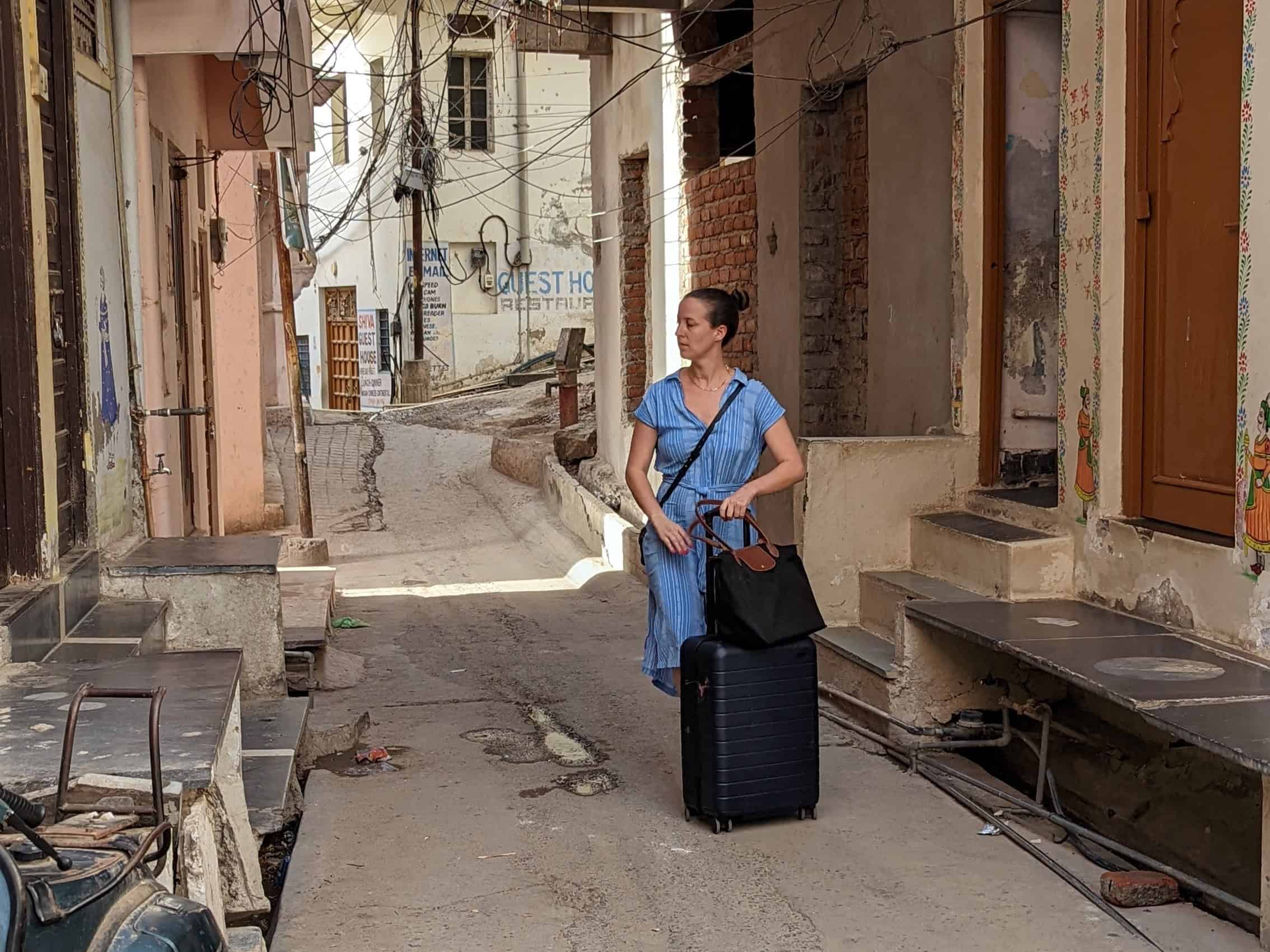 A girl in a light blue dress with short sleeves pushing a rolling luggage through a small empty street in Udaipur.