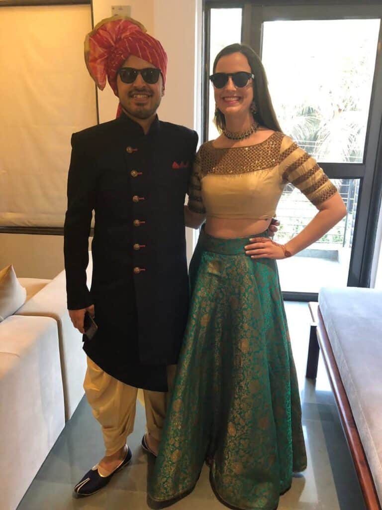 Young man and woman dressed in traditional Indian wear inside a beige room. The man is wearing a navy sherwani with a red turban and the woman is in a gold blouse and green and gold lehenga. They are both wearing sunglasses.