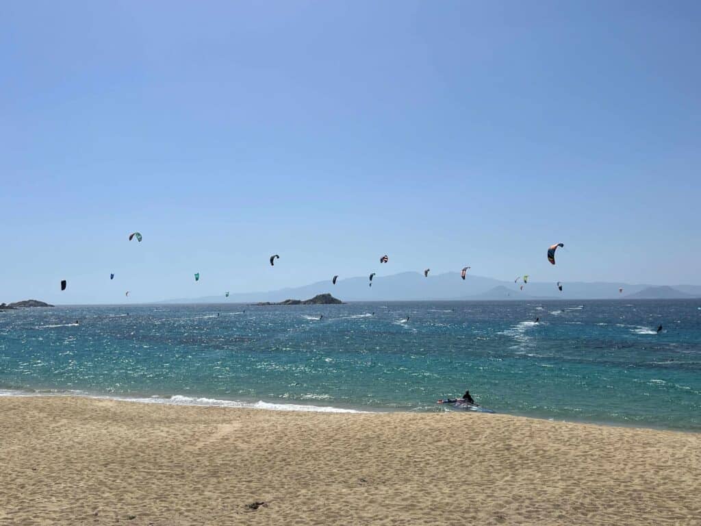 View of Mikri Vigla Beach on Naxos Island, Greece, with several kite surfers riding the waves under a clear blue sky. Colorful kites are scattered across the horizon, while the turquoise sea contrasts with the golden sandy beach in the foreground. Distant hills and small islands can be seen in the background.