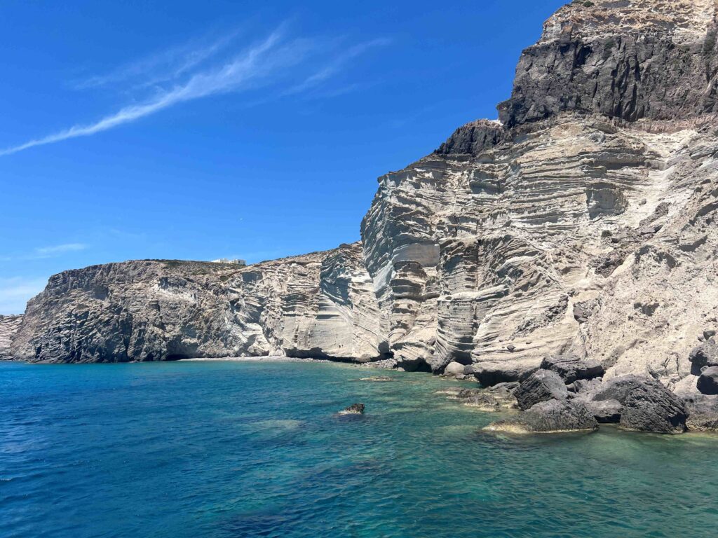 Rocky cliffs with layered textures rise steeply from the turquoise-blue waters off the coast of Paros, Greece. The rugged white and gray rock formations create a dramatic contrast against the clear blue sky above. Small patches of greenery can be seen on top of the cliffs.