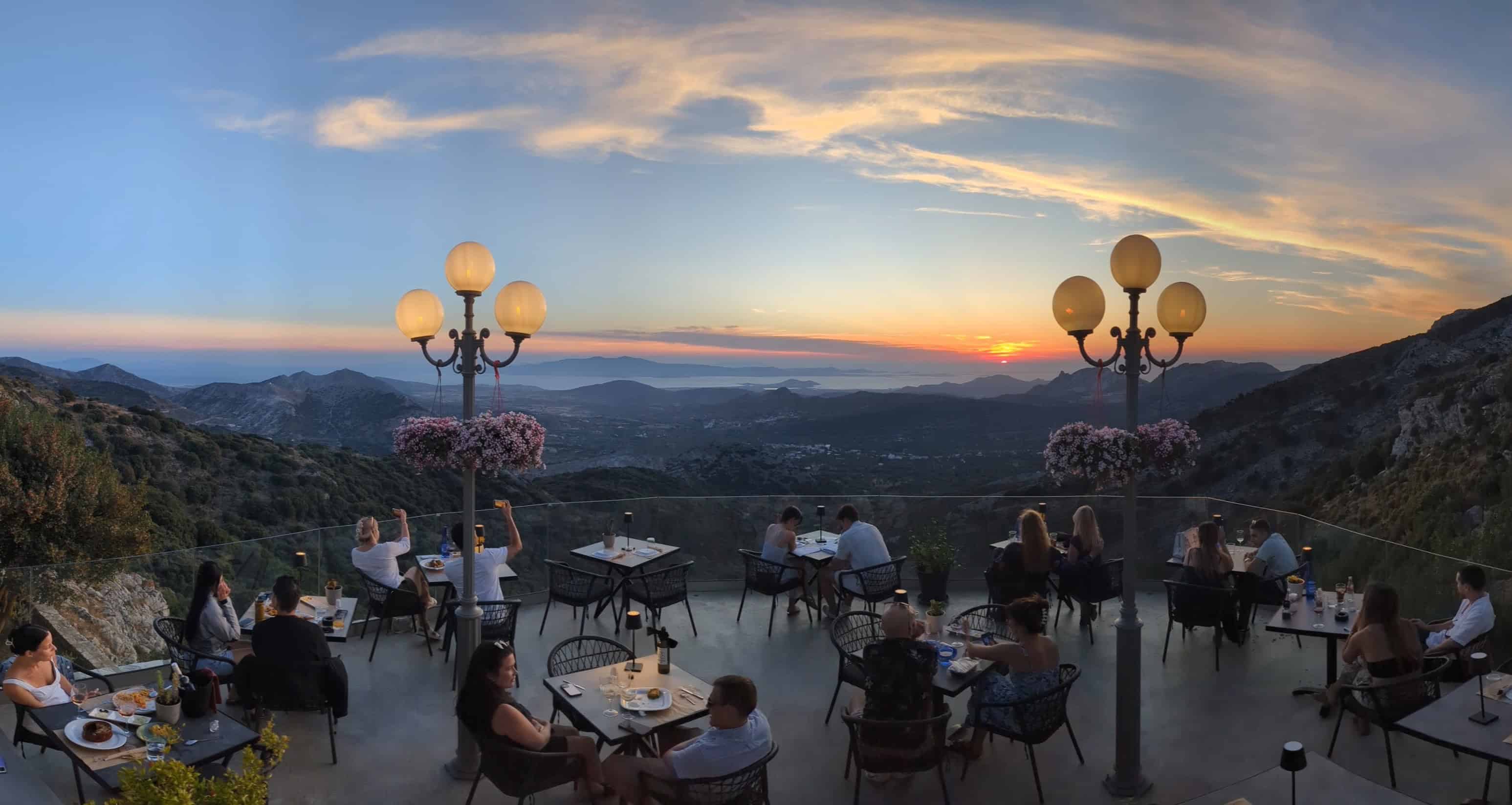 Terrace dining at Rotunda Restaurant in Naxos with a handful of couples at tables and the sunset over the mountains and sea in the distance.