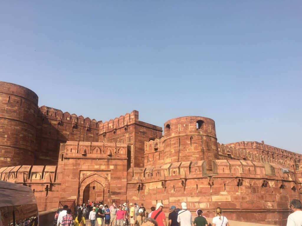 View of the red sandstone walls of Agra Fort against a blue sky with people walking towards an open archway to enter.