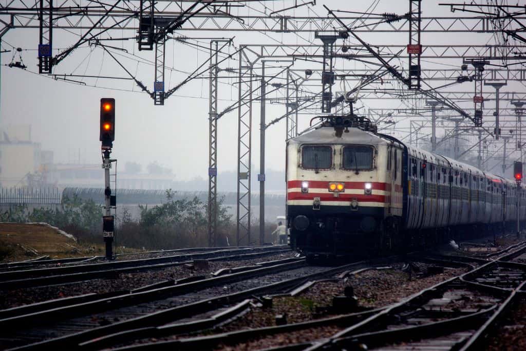 An Indian Railways train approaches through a network of tracks and overhead wires on a misty day, with signals glowing red in the foreground.