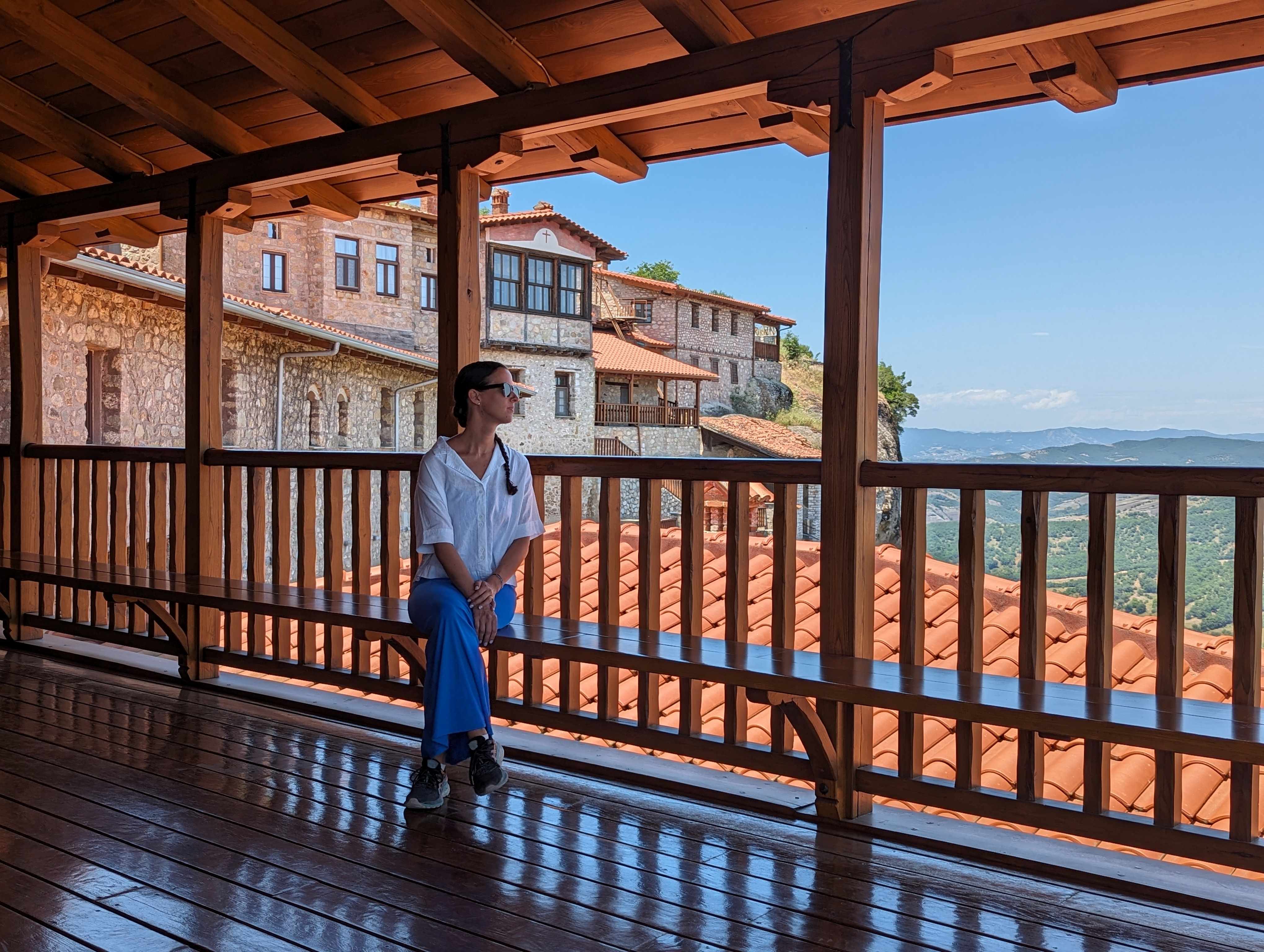 A woman sits on a bench inside the Holy Monastery of the Great Meteoron looking out on the view below of green valleys and blue sky.