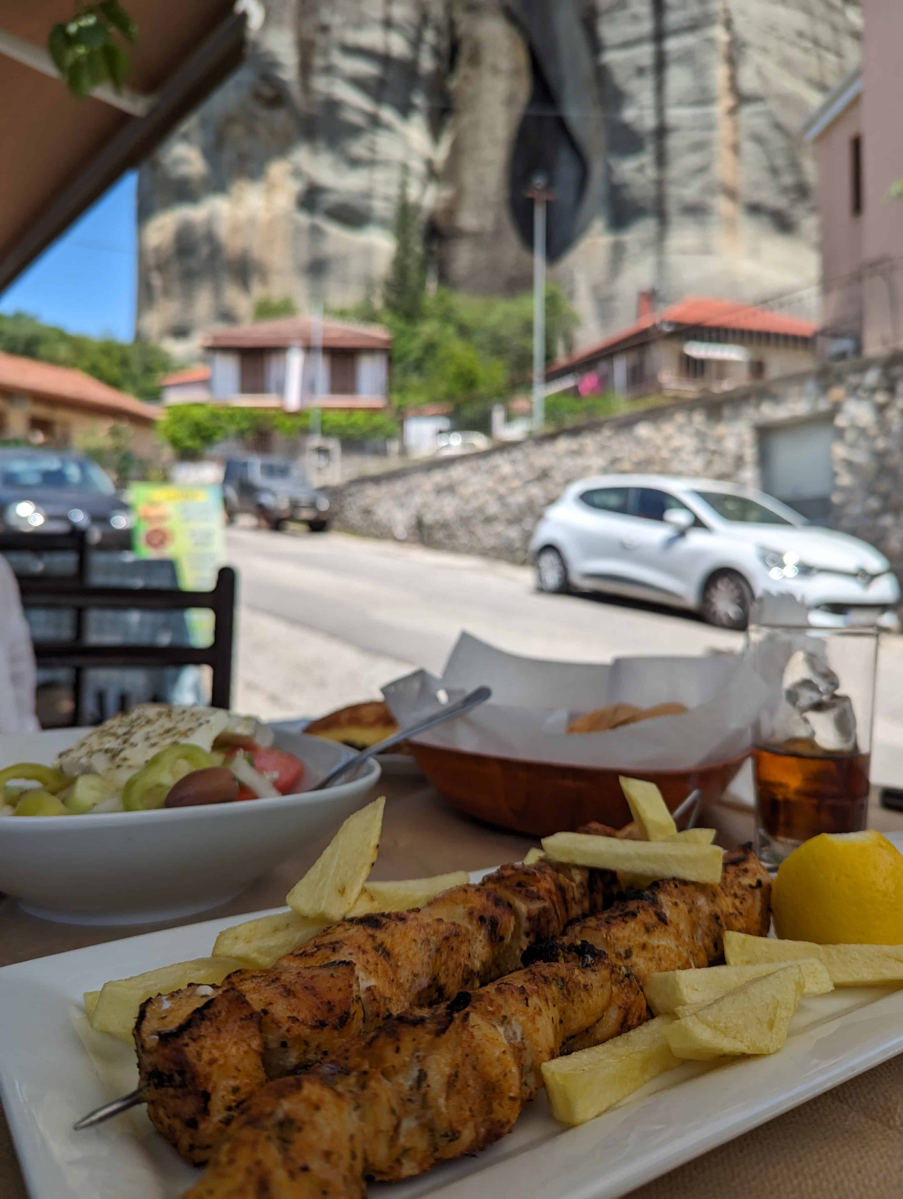 A plate of grilled chicken skewers with potatoes and a side of Greek salad, served at a taverna with a view of a rocky mountainside and a small village street in Meteora, Greece.