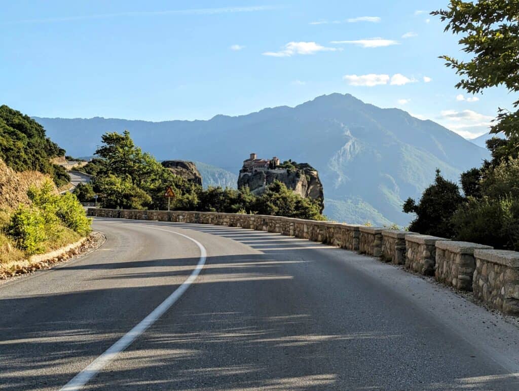 A winding road leads towards a majestic monastery perched atop a towering rock formation in Meteora, Greece. The road is surrounded by lush greenery and mountains, with a clear blue sky above.