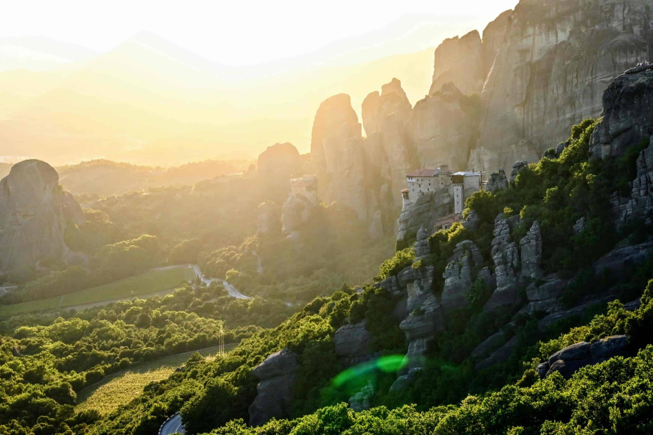 A breathtaking panoramic view of Meteora, Greece. The image showcases towering, sandstone rock formations with ancient monasteries perched atop them. Lush greenery covers the valley below, and the setting sun casts a warm, golden glow over the landscape.