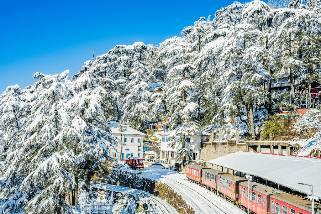 Snow-covered pine trees surround a quaint railway station in a serene mountain town, with a bright red train standing still on the tracks. The landscape is blanketed in fresh snow, creating a picturesque winter scene against a clear blue sky.