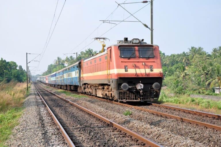 An Indian Railways train traveling through a lush, green countryside, showcasing the iconic red locomotive and blue passenger cars. The train is moving along a track under a clear sky, surrounded by trees and rural scenery.