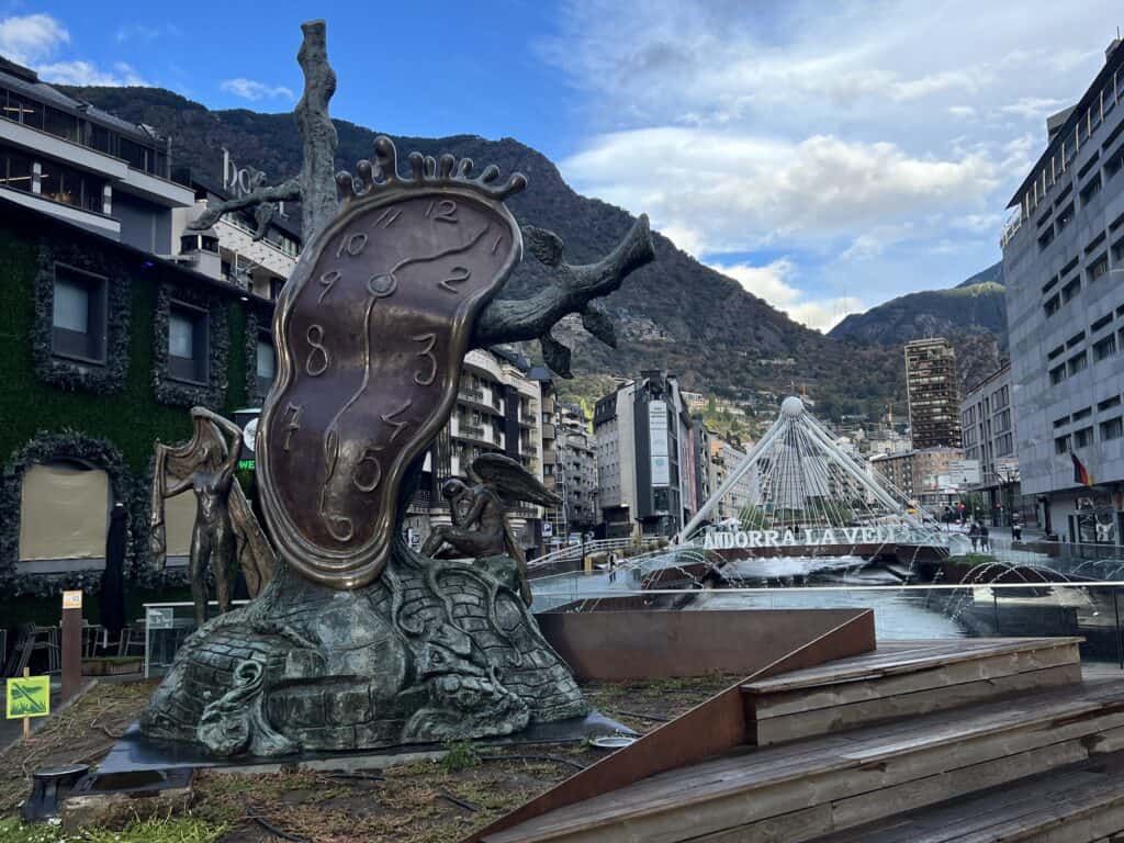 Salvador Dalí's iconic melting clock sculpture in the heart of Andorra la Vella, Andorra. The bronze artwork, known as 'La Noblesse du Temps,' stands prominently near the city's modern footbridge. Surrounded by buildings and dramatic mountain scenery, the artwork reflects Dalí’s surrealist style, blending with the urban landscape against a backdrop of blue sky and rugged peaks.