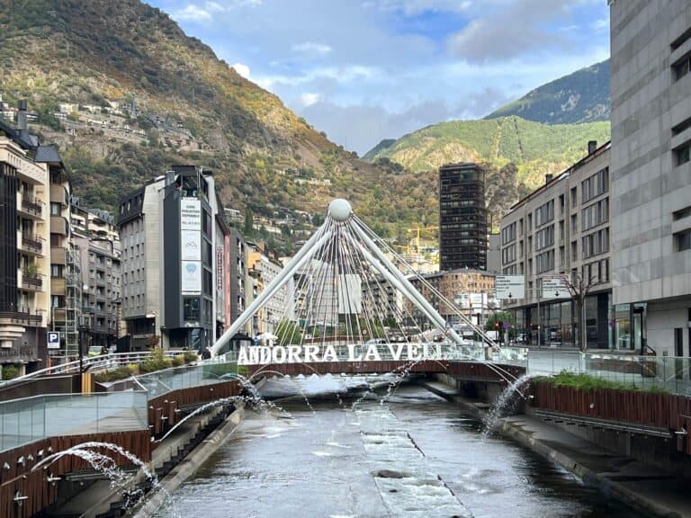 A view of the iconic bridge in Andorra la Vella, spanning a river with mountain landscapes in the background. The bridge features the city's name, "Andorra la Vella," in large white letters. Modern buildings and shops line the streets on both sides, while fountains spray water into the river, adding a dynamic element to the scene. The lush, green mountains rise behind the city, showcasing the blend of nature and urban life in Andorra's capital.