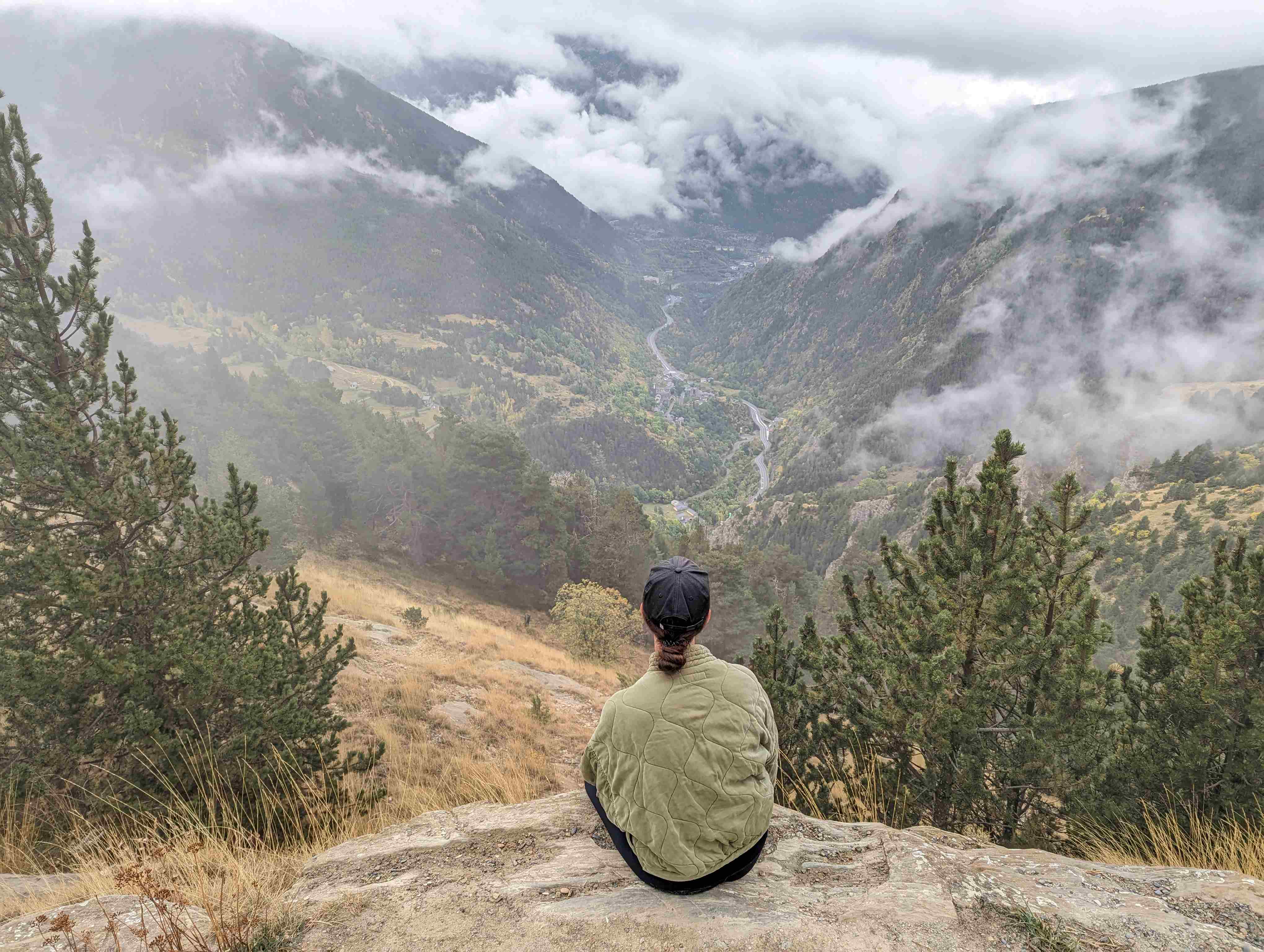 A girl wearing a green quilted jacket and black cap sits on a rocky ledge overlooking a misty valley in Andorra. The view features rolling mountains shrouded in low-hanging clouds, a winding road cutting through the landscape, and a mix of pine trees and golden grasses, creating a serene and contemplative scene.