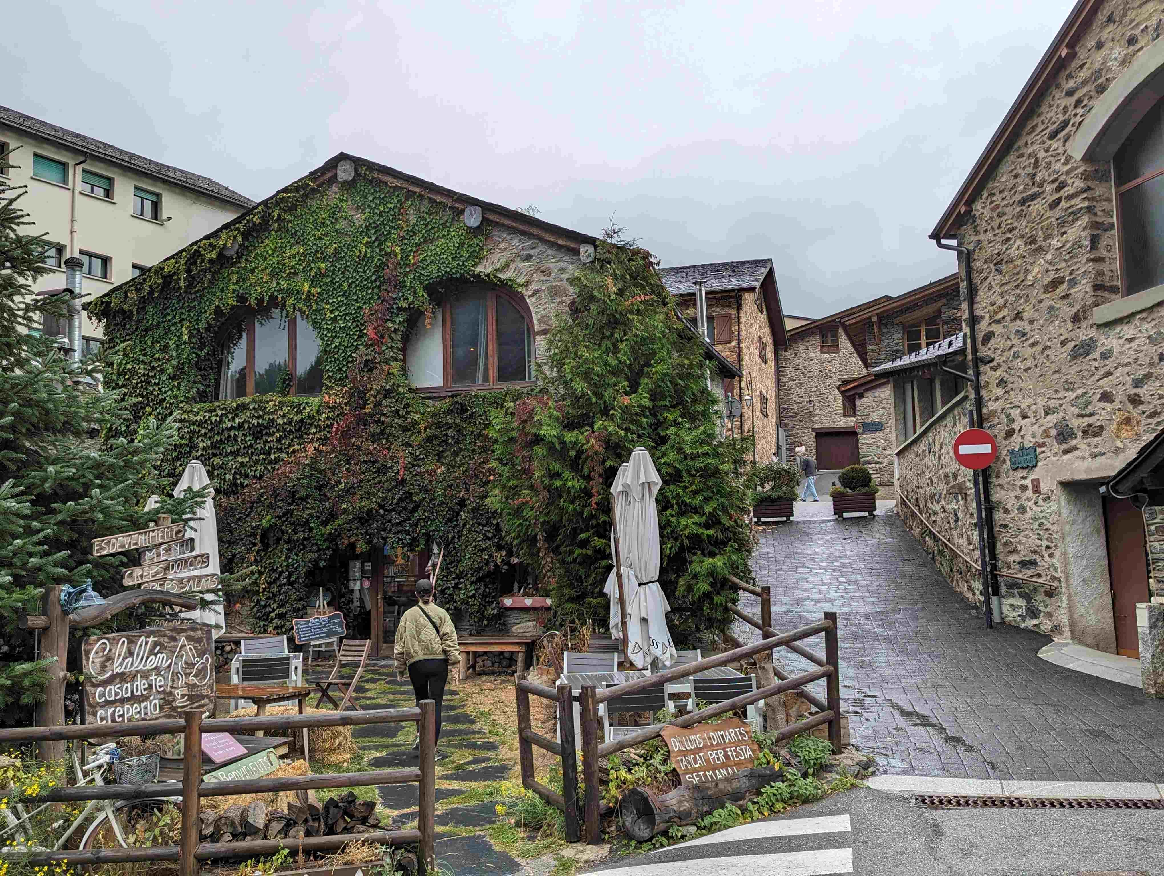 A charming stone building covered in ivy in Ordino, with arched windows and rustic character. The façade is partially covered in green climbing plants. In front is a casual café or creperie with wooden fence railings, outdoor seating, and handwritten signs. A cobblestone path leads uphill past other traditional stone buildings. The scene captures the quaint, historic atmosphere of this Andorran mountain village.