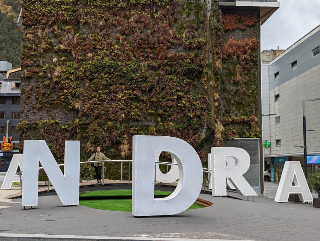 A person standing inside the large white "ANDORRA" letters installation, positioned against a vertical green wall covered with plants and autumnal foliage. The scene is set in a modern urban area with surrounding buildings, capturing a mix of nature and city vibes.