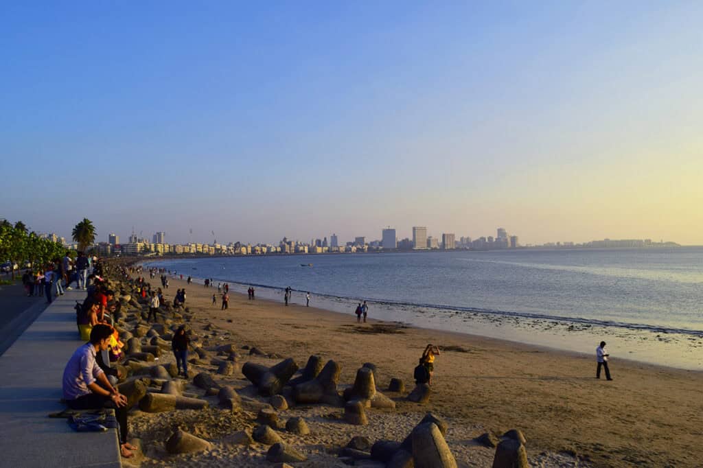 People sitting along a sea wall with a sandy beach and seaside views, enjoying a sunset at the picturesque Marine Drive in Mumbai, India. You can see some iconic buildings in Mumbai and the South Mumbai skyline in the background.