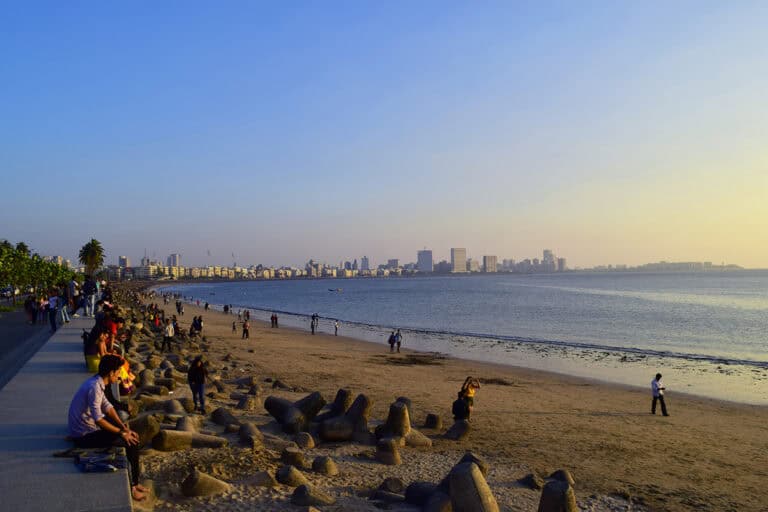 People sitting along a sea wall with a sandy beach and seaside views, enjoying a sunset at the picturesque Marine Drive in Mumbai, India. You can see some iconic buildings in Mumbai and the South Mumbai skyline in the background.