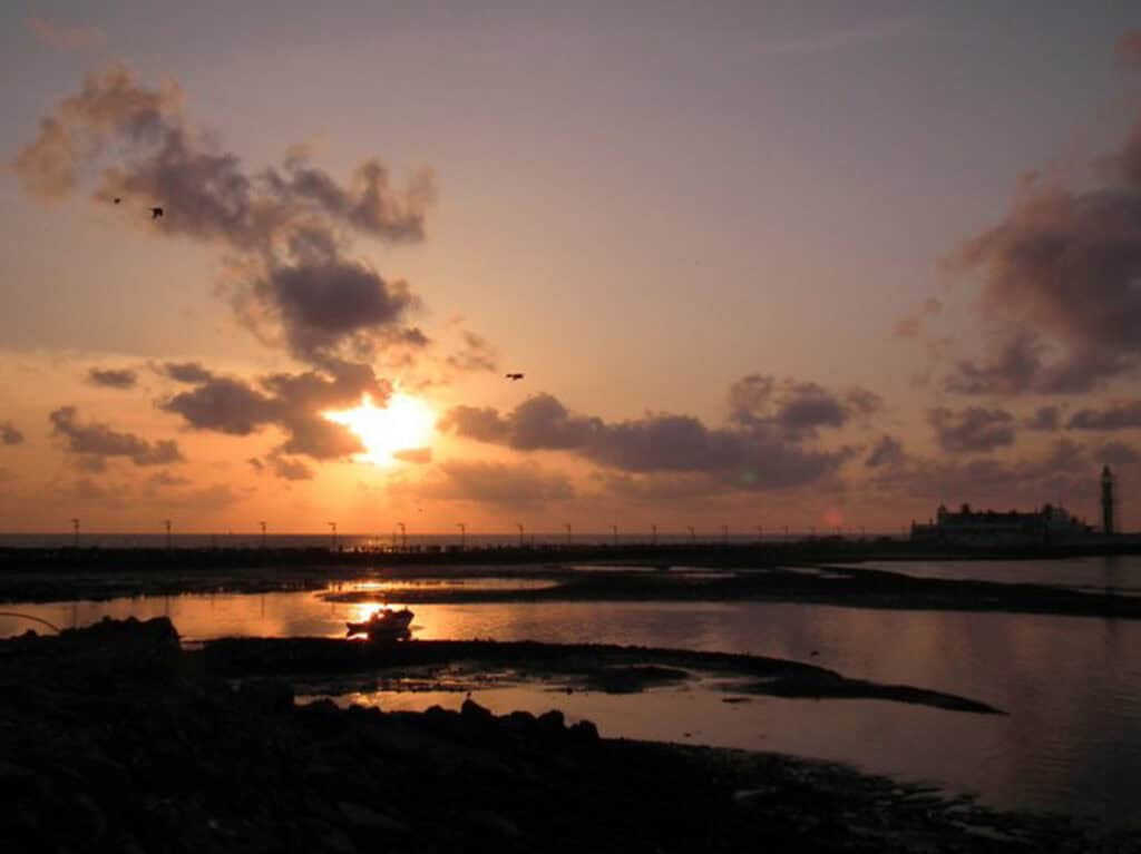 A sunset view of the Haji Ali Mosque in Mumbai, India. There is a mosque in the sea with a walking platform that runs through the sea towards it. The shot is taken at low tide and there is a boat along with coastal rocks that can be seen.