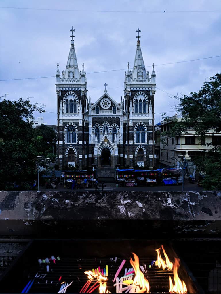 The stone built Mount Mary Basilica in Mumbai, Bandra which is a church from the 1600s. It is built with black stone and the picture shows two church towers and candles burning outside on a stall.  