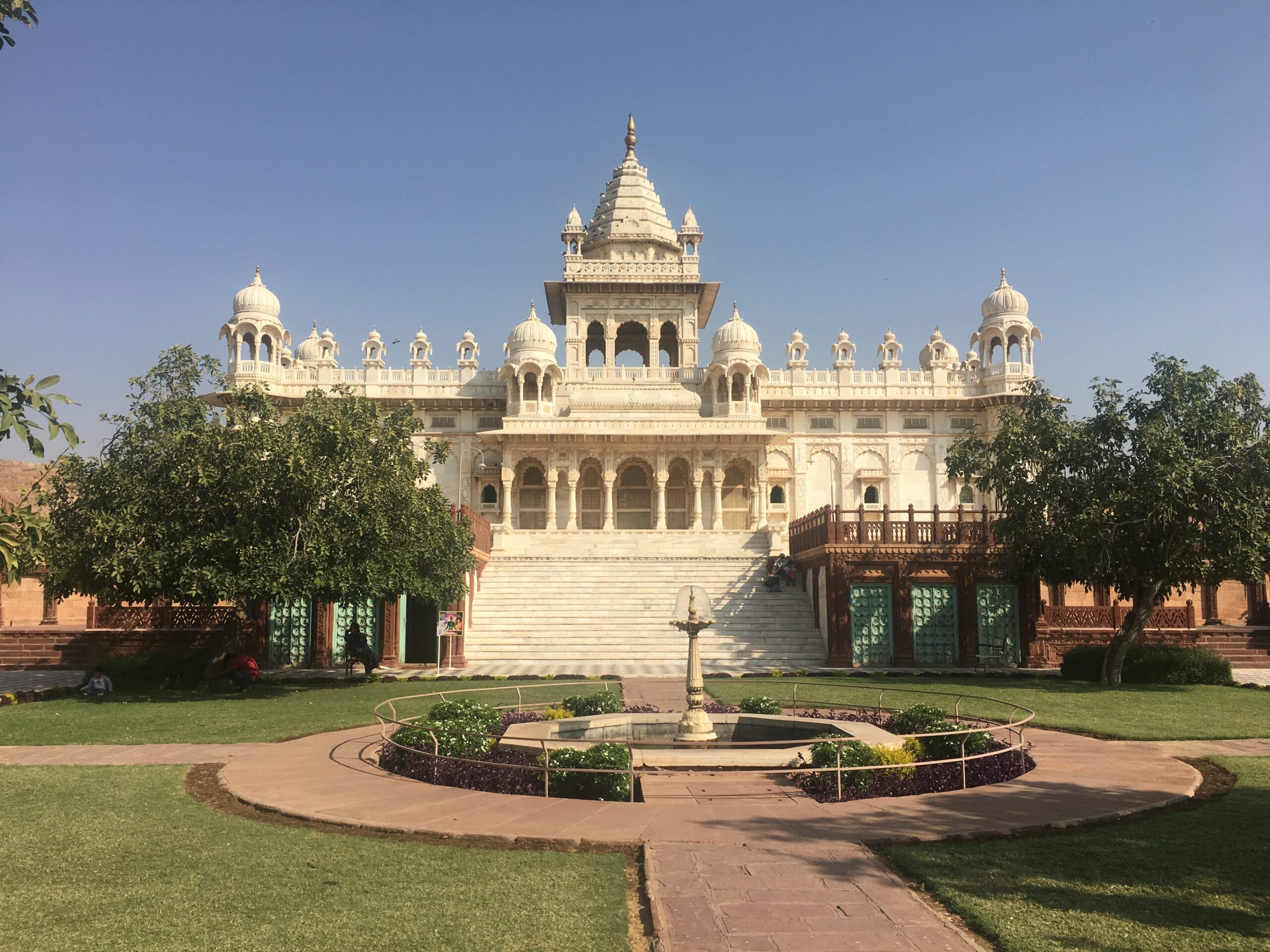 A grand white marble structure with intricate domes and arches, standing against a clear blue sky. The building, set amidst a well-manicured garden with a central fountain, features ornate architecture typical of royal Rajasthani palaces. 