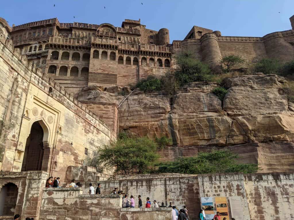 isitors walking towards the grand entrance of Mehrangarh Fort in Jodhpur, India, with the towering fort walls and intricately carved architecture rising dramatically above the rocky landscape.