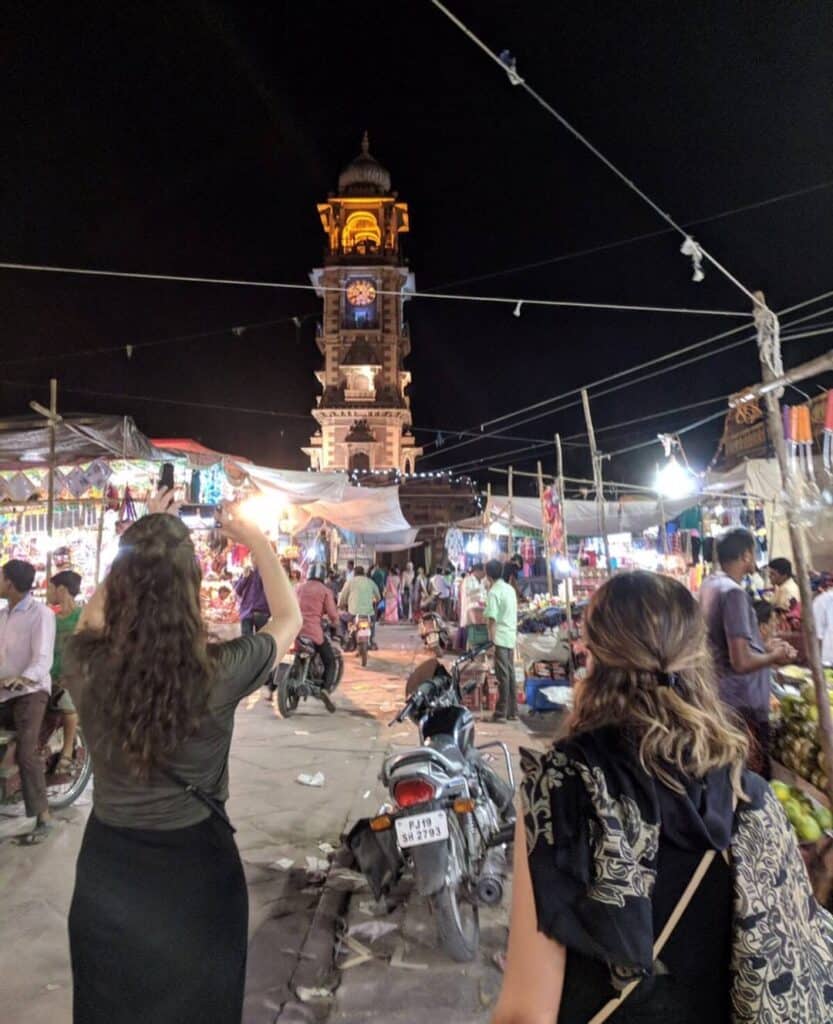 A bustling night market in Jodhpur with shoppers walking through stalls filled with colorful goods. The iconic Clock Tower, brightly illuminated, stands prominently in the background. Two women in the foreground capture the lively scene on their phones, while motorcycles and pedestrians navigate the busy market area