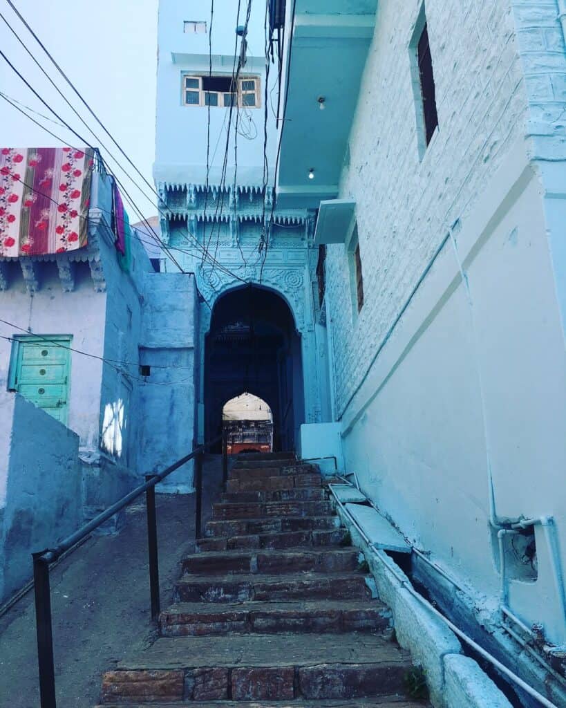 A narrow stone staircase leads up through a passageway between light blue-painted buildings in Jodhpur. The walls are adorned with intricate architectural details, and colorful fabrics hang from the balconies above. Wires crisscross overhead, adding to the rustic charm of the old city streets. The alleyway offers a glimpse into Jodhpur's famous blue city architecture