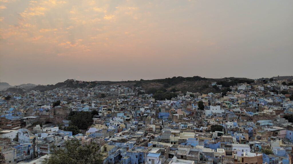 A view from Pachetia Hill in Jodhpur at sunset, overlooking the city's famous blue buildings with the sun setting behind distant hills, casting a warm glow across the sky.