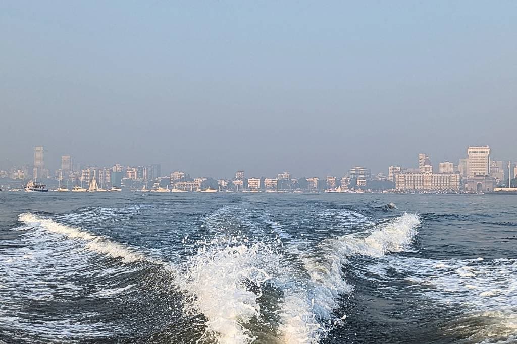 View of Mumbai’s skyline from the water, showing buildings along the coastline and boats on the sea. The photo captures the wake of a boat in the foreground, with calm waters leading toward the cityscape, partially shrouded in haze under a soft blue sky. Notable landmarks, including the Gateway of India, are visible along the shoreline.