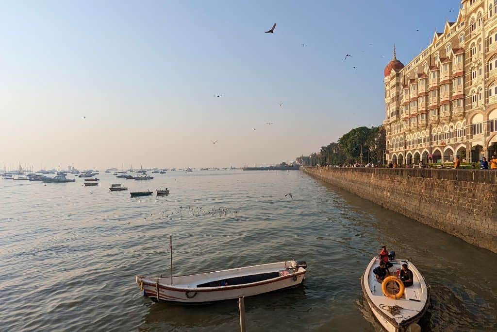 View of Mumbai’s waterfront with boats floating on the calm Arabian Sea meeting the sea wall. The iconic Taj Mahal Palace Hotel stands on the right, its distinctive dome and architectural details in the golden light of the late afternoon. Small boats are docked in the foreground, while birds soar above the water, and the horizon stretches into the hazy distance with more boats scattered.