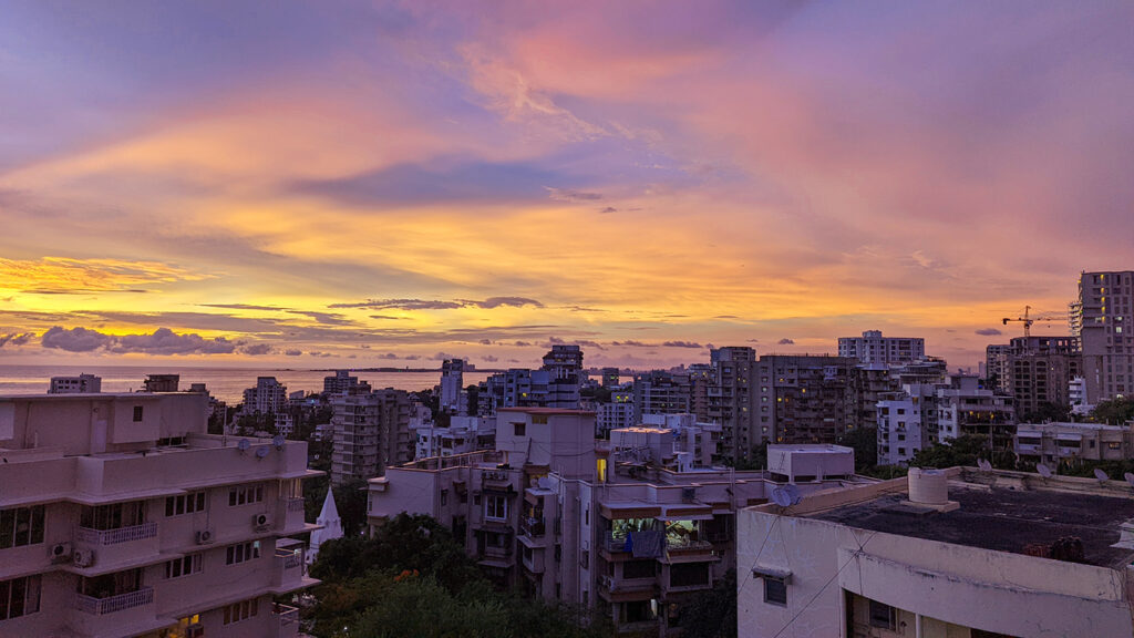 A summer sunset in Bandra, Mumbai. There are shades of blue, yellow, orange and pink in the cloudy sky. You can see the suburban buildings of Bandra in the foreground with the sea and the sky in the background. 