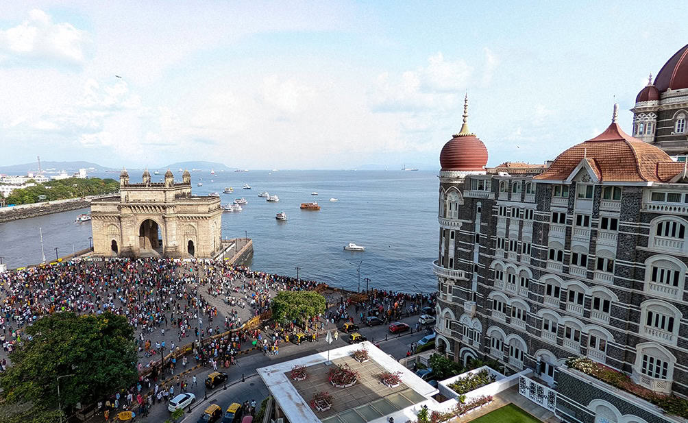 A view of the Gateway of India arch by the Arabian Sea from the Taj Mahal Palace Hotel in Mumbai. There is a crowd of people gathered by the seaside arch across the street with a view of the ornate hotel on the right.