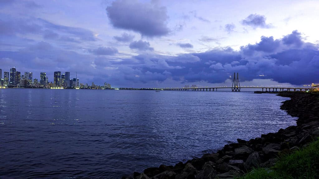 An evening view of the Mumbai skyline over the bay. You can see the well lit Bandra-Worli sea link spread across the bay with sky scrapers on the left. There are ominous clouds hanging over the bay.