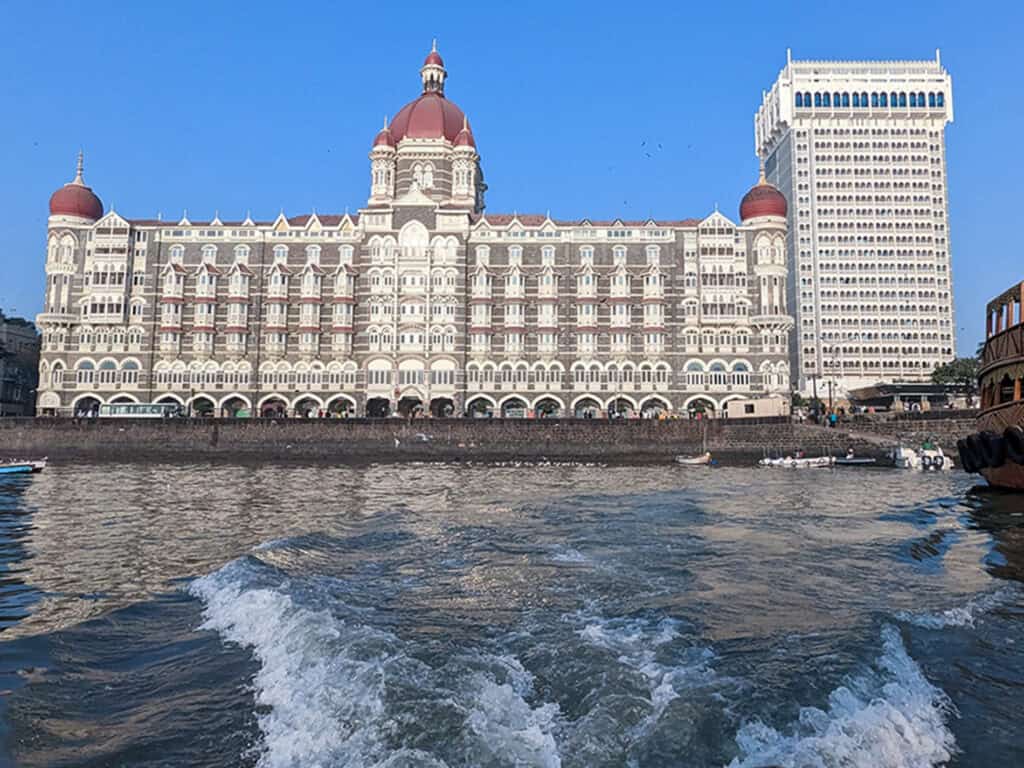 A view of the historic, ornate Taj Mahal Palace Hotel in Colaba Mumbai from a boat in the sea. There are people along the promenade with the hotel in the background. 