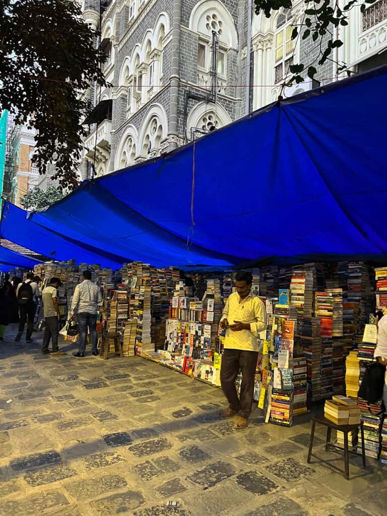 Street side book stalls with piles of books under a blue tarp in the historic Fort district of Mumbai. There are book sellers by the stall and you can also a typical gothic structure in South Mumbai. 