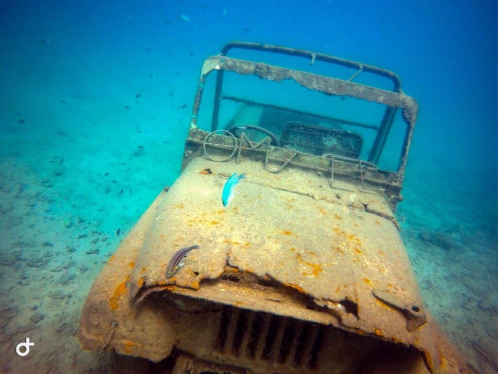 An underwater picture of a jeep that is on the sea bed. The water is clear blue and emerald and there are two fishes in the frame. The jeep is rusty. 