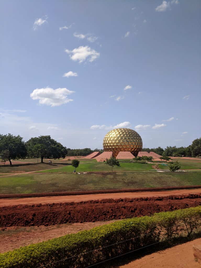 A picture of the golden dome temple in Auroville, India. It is a golden metal dome in the centre of a large garden. 