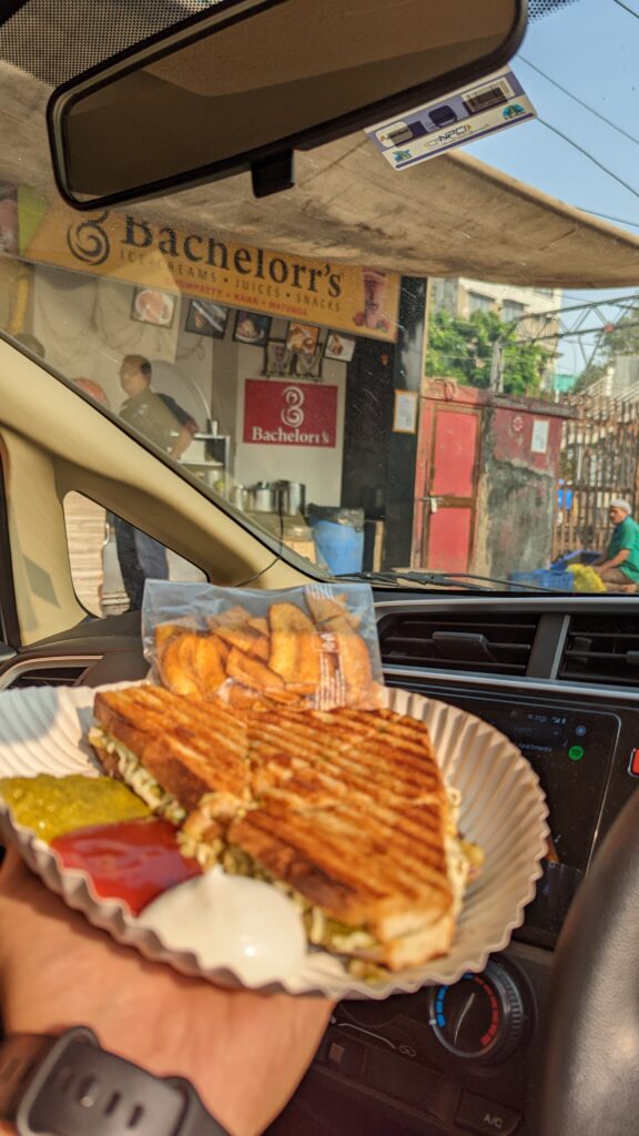 A picture of a unique Bombay street sandwich with banana chips. There is ketchup and mayo on a paper plate inside a car. 