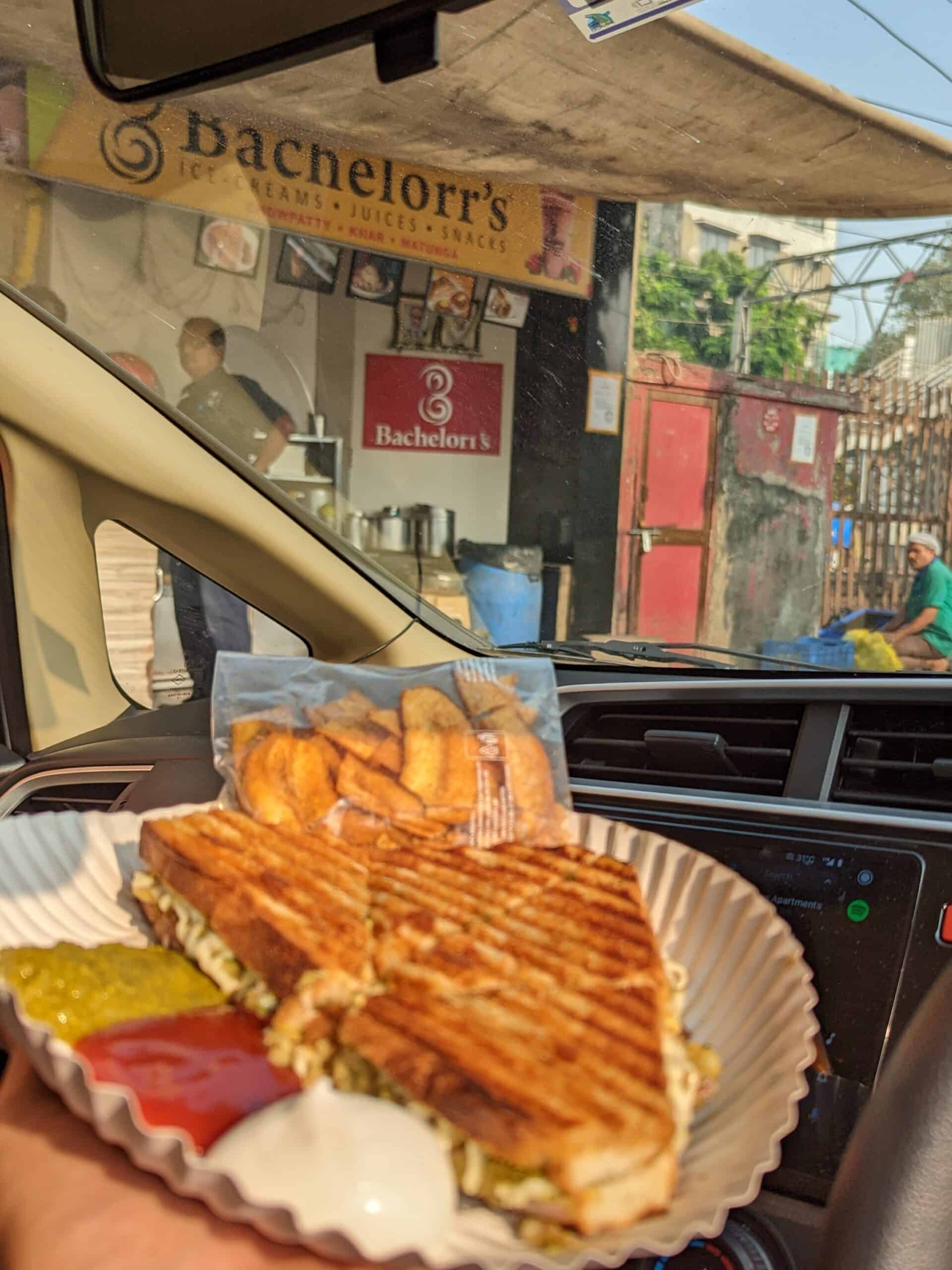 A picture of a unique Bombay street sandwich with banana chips. There is ketchup and mayo on a paper plate inside a car.