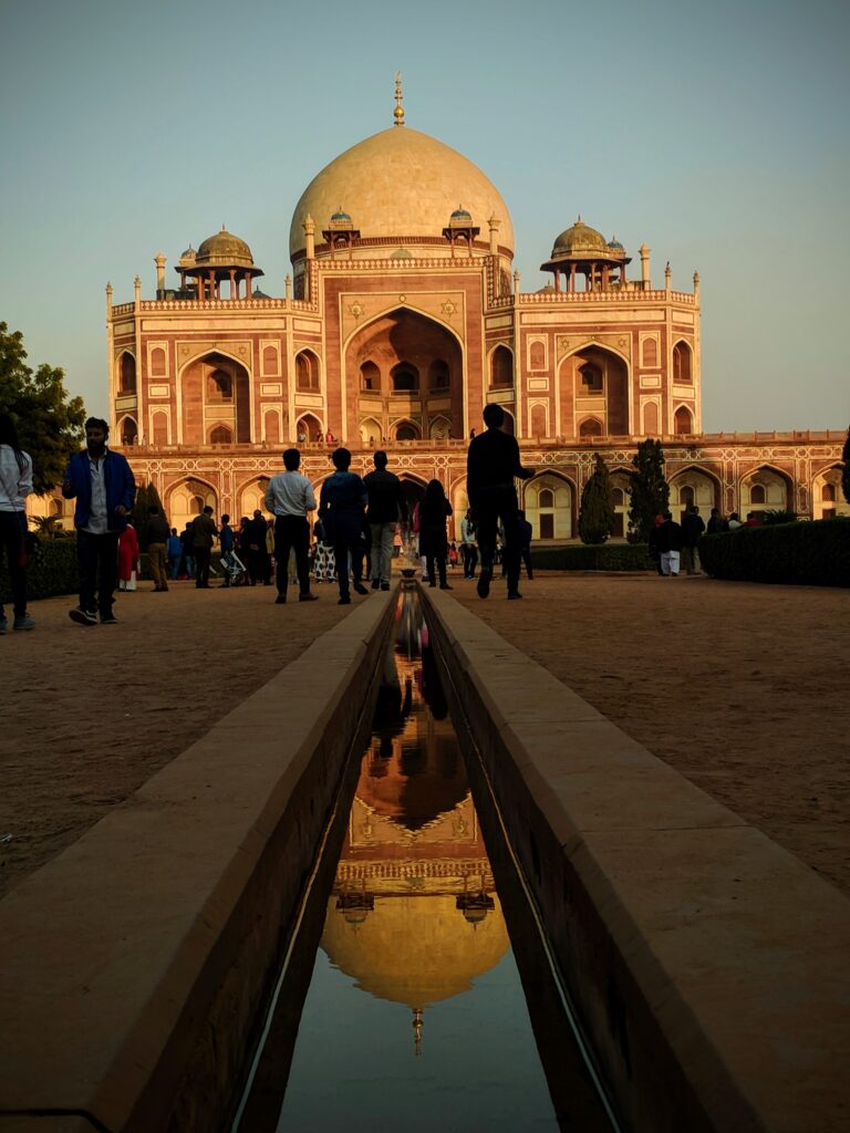 A picture of the Humayun Tomb monument in Delhi. The picture is taken from ground level looking up at the red sandstone and marble monument. There are 3 domes with one prominent dome in a medieval Islamic style architecture. The main dome is reflecting in a water fountain in the foreground. There is a red, sunset hue to the picture. 